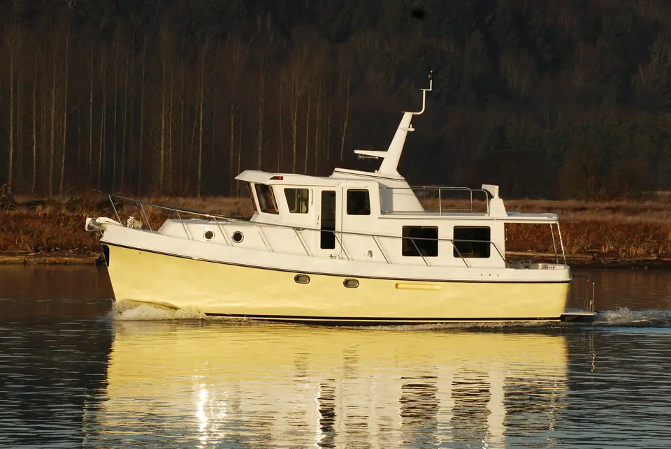 American Tug 435 with a cream hull cruising in profile past a wooded shoreline