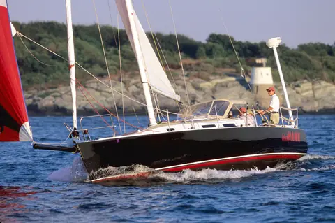 Dark hulled J/120 sailing near a lighthouse as a red spinnaker is hoisted
