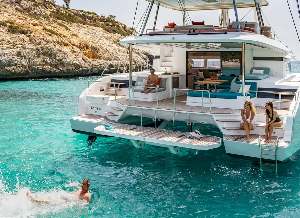 Lagoon 55 at anchor in a turquoise cove seen from astern with swimmers off the transom steps