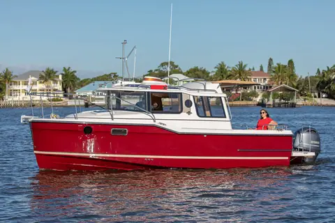 Ranger Tugs R-23 with a red hull and outboard cruising in profile past a palm lined shore