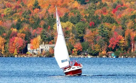 Red hulled Rhodes 22 heeling under sail past autumn foliage on a lake
