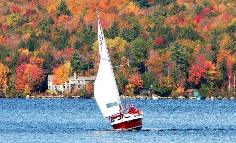 Red hulled Rhodes 22 heeling under sail past autumn foliage on a lake