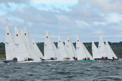 Fleet of Shields one design keelboats racing upwind in a breeze with the red tulip on their mainsails